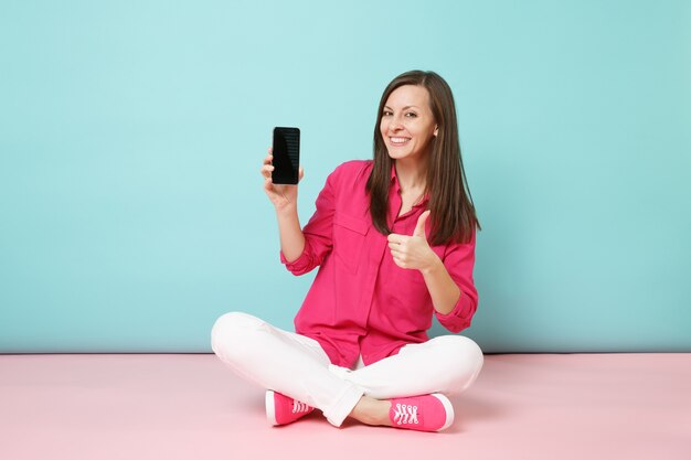 Full length portrait of young fun woman in rose shirt, white pants sit on floor hold cellphone 