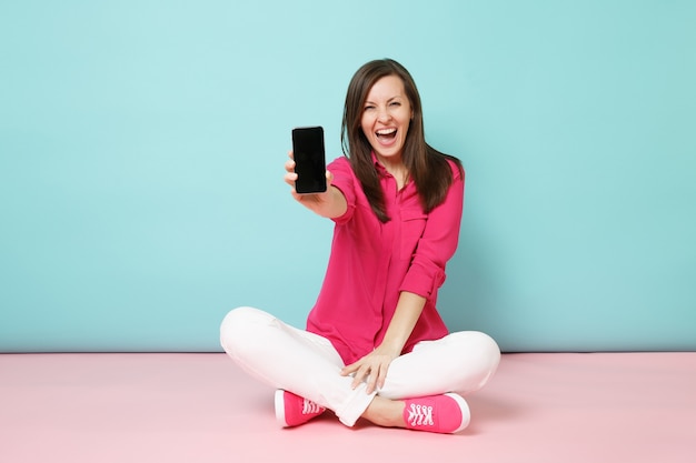 Full length portrait of young fun woman in rose shirt, white pants sit on floor hold cellphone 