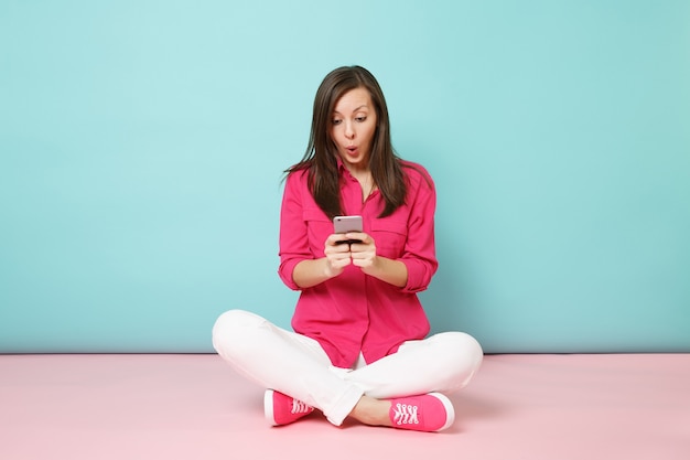 Full length portrait of young fun woman in rose shirt, white pants sit on floor hold cellphone 