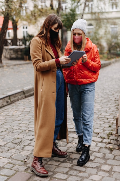 Full length portrait of young ladies dressed in warm clothing holding digital tablet while standing on street. Happy woman wearing medical protective masks during quarantine.