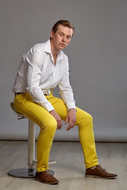 Full length portrait of a young lovely ginger person in a stylish white shirt and yellow trousers looking at the camera while posing sitting sideways on a bar chair against a gray studio background. H