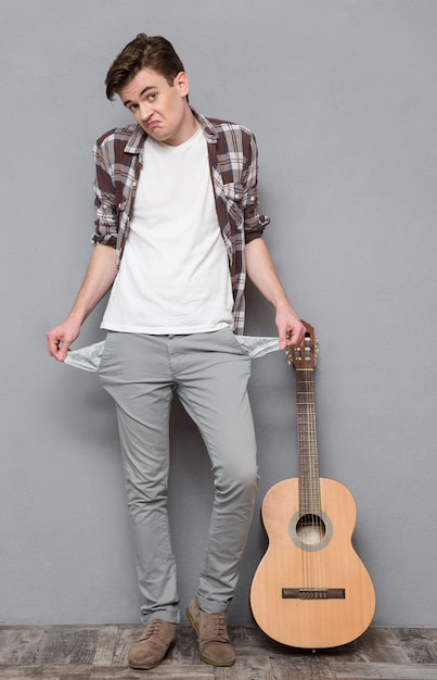 Full length portrait of a young man exhibiting his empty pockets on gray bakground