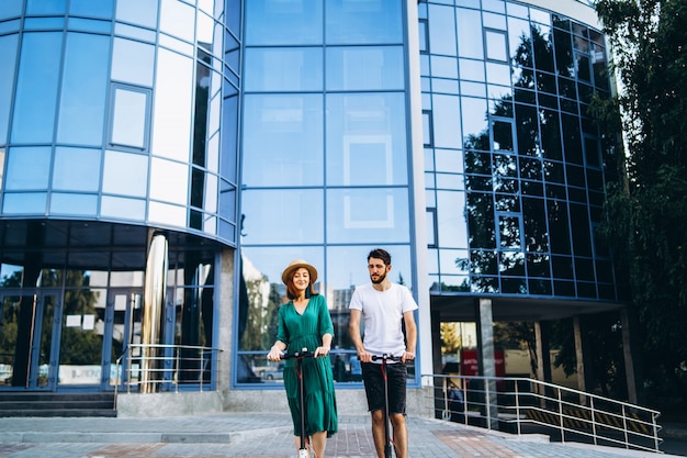 Full length portrait of a young romantic couple with electric scooters, walking in the city. Young woman in hat and man enjoy a walk