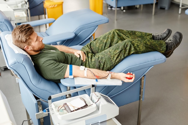 Full length portrait of young soldier donating blood while lying in comfortable chair at med center