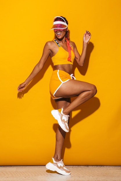 Полный портрет молодой стильной африканской женщины в спортивной одежде, стоящей изолированно над желтой стеной и позирующей