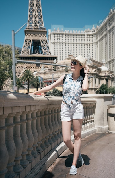 Photo full length portrait of young stylish woman traveler in hat near eiffel tower in paris on sunny day. smiling girl tourist standing on hot summer outdoor. joyful female backpacker leaning on railing.