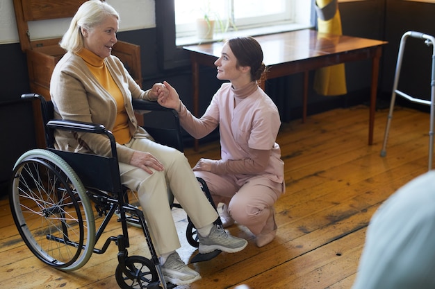 Full length portrait of young woman assisting smiling senior woman in wheelchair at nursing home cop...