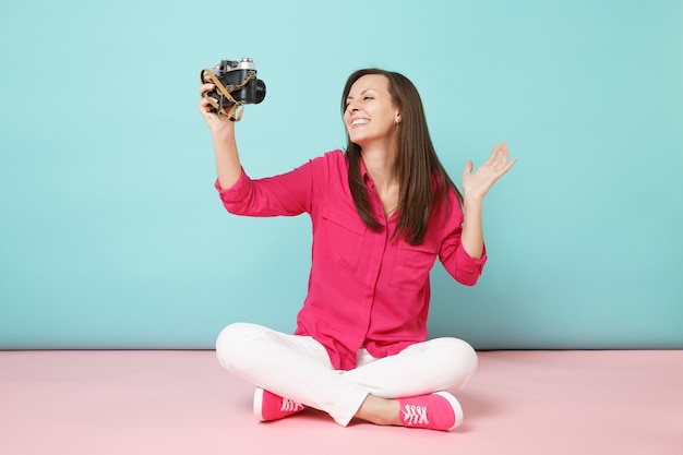 Full length portrait young woman in rose shirt blouse white pants sitting on floor hold camera 