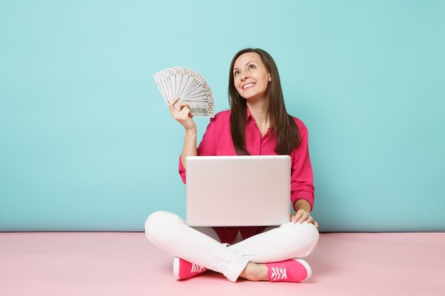 Full length portrait of young woman in rose shirt blouse, white pants sitting on floor hold money pc 