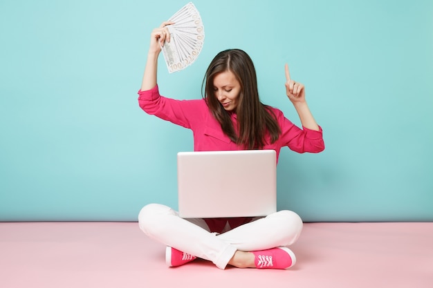 Full length portrait of young woman in rose shirt blouse, white pants sitting on floor hold money pc 