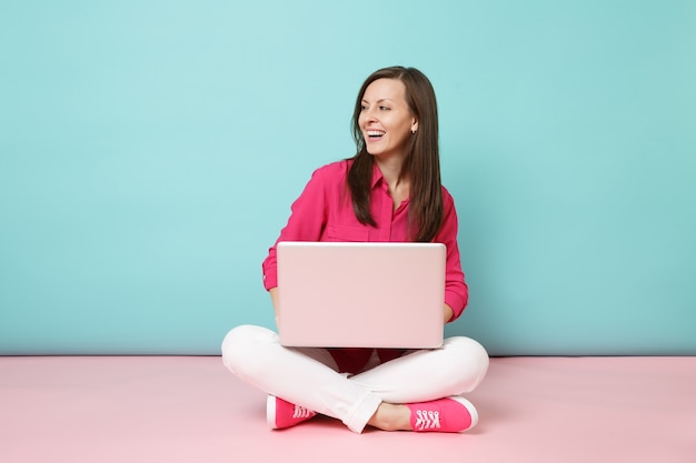 Full length portrait of young woman in rose shirt blouse white pants sitting on floor using laptop pc 