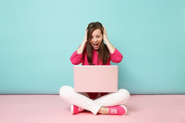 Full length portrait of young woman in rose shirt blouse white pants sitting on floor using laptop pc 