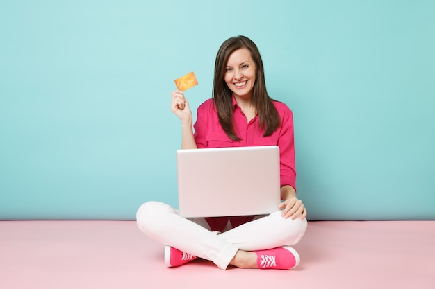 Full length portrait of young woman in rose shirt, white pants sitting on floor hold credit card pc 