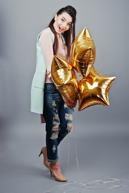 Full-length portret jonge brunette meisje draagt in roze blouse, turquoise jas, gescheurde spijkerbroek en crème schoenen met gouden ster ballonnen. Mode studio-opname