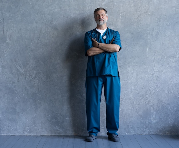 Full-length portret van professionele volwassen chirurg die naar de camera kijkt terwijl hij tegen de grijze achtergrond staat.