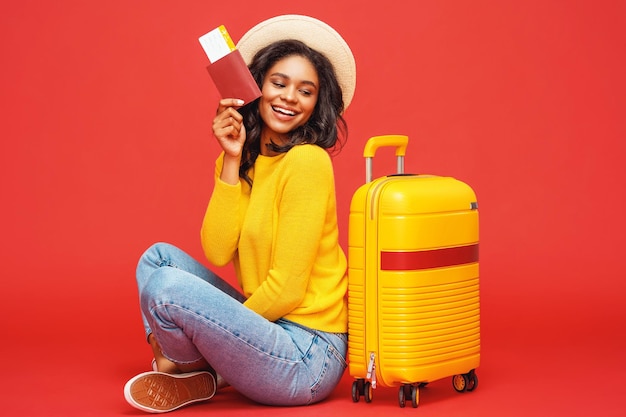 Photo full length positive ethnic tourist with passport and tickets smiling and dreaming about vacation while hugging suitcase against red backdrop