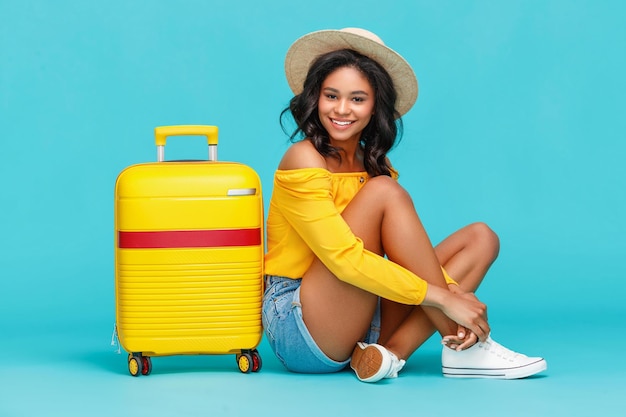 Full length positive ethnic woman with hat smiling for camera while sitting cross legged near suitcase during summer vacation against turquoise background