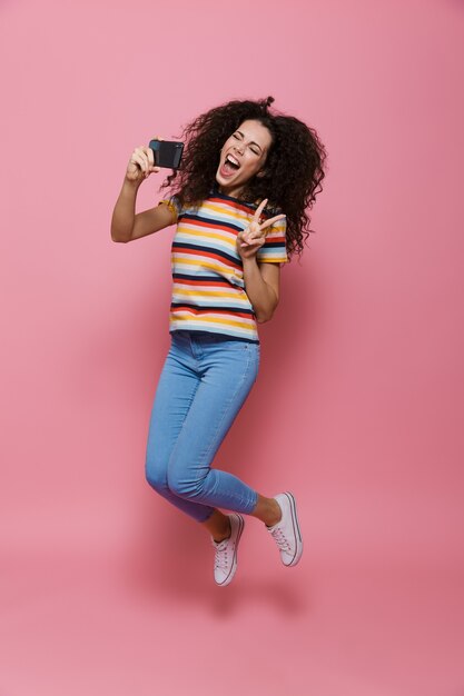 Full length of positive woman 20s with curly hair holding cell phone and taking selfie photo isolated on pink