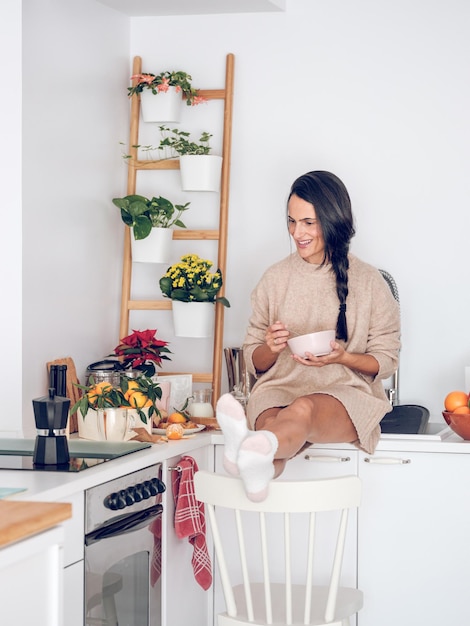 Full length of positive young Hispanic woman with long dark hair in casual clothes and socks smiling while sitting on kitchen counter with legs on chair and having breakfast