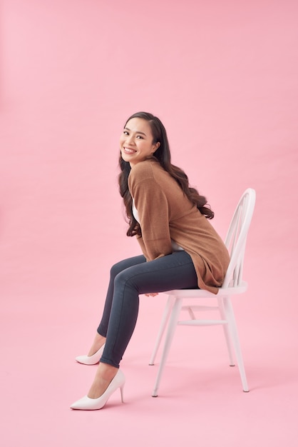 Full length of a pretty casual woman sitting and looking at camera isolated on the pink background