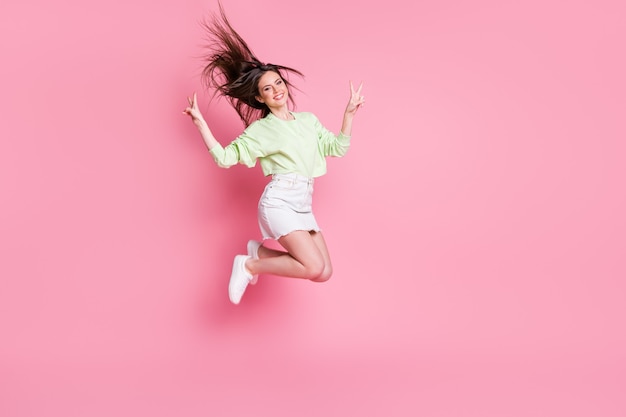 Full length profile photo of attractive youngster lady jumping high showing v-sign symbols wear casual crop pullover naked belly skirt shoes isolated pink color background