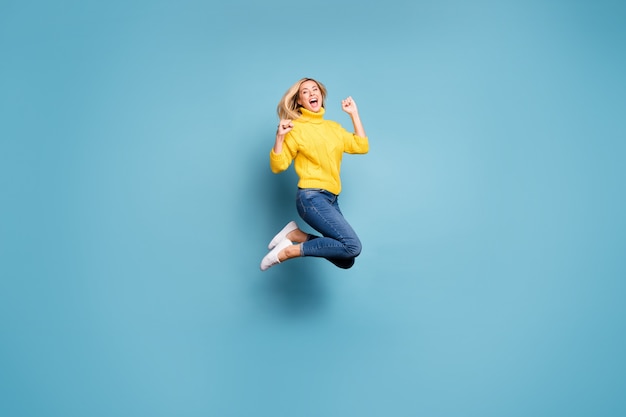 クレイジーな女性が高くジャンプして海外への無料旅行の勝利を祝うフルレングスのプロフィール写真は、ニットの黄色のプルオーバージーンズを分離した青い色の壁を喜んで着用します