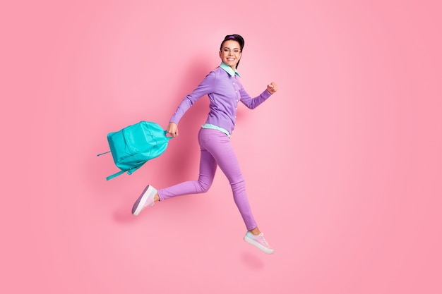 Full length profile photo of girl hold backpack jump go look camera wear specs purple jumper pants sneakers isolated pink color background