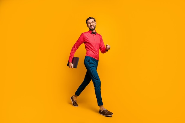 Full length profile photo of macho business man walk corporate meeting training carry laptop wear stylish red shirt bow tie blue pants shoes 