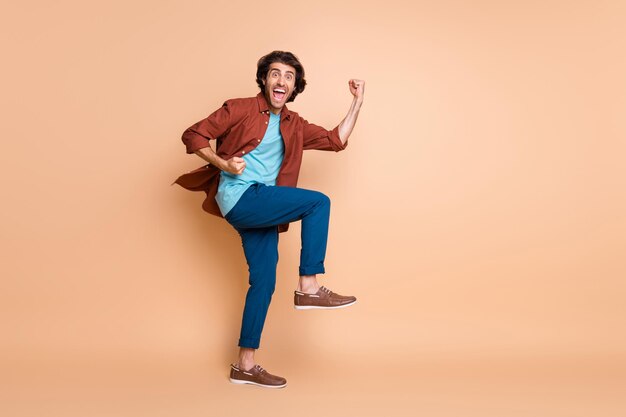 Full length profile photo of man raise fists open mouth look camera wear brown t-shirt blue pants shoes isolated beige color background