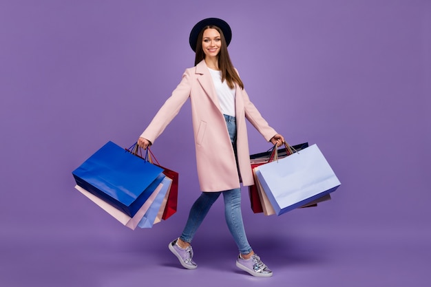 Photo full length profile side photo of cheerful lovely trendy girl hold many bags go from shopping mall wear beige outfit denim headwear sneakers isolated over purple color background