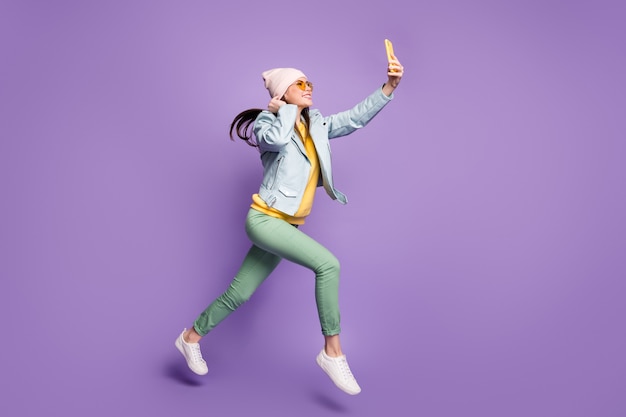 Полная длина профиля сбоку фото симпатичной довольно милой девичьей девушки прыгать, бегать, вести блог, делать селфи, смартфон, видеозвонок, носить стиль, зеленые брюки, брюки, изолированные на фиолетовом цветном фоне