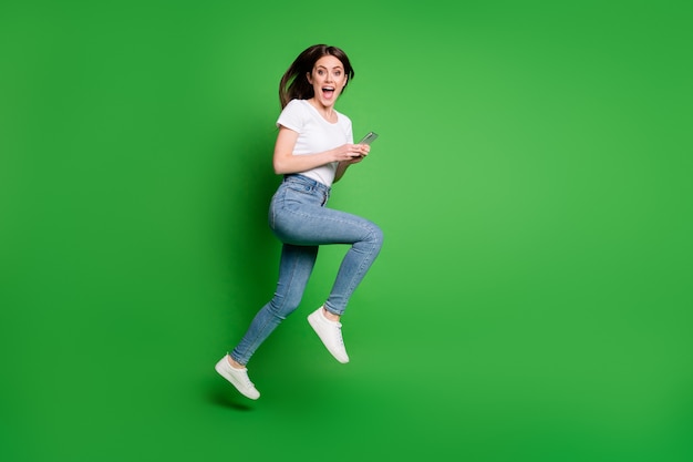女の子のジャンプランのフルレングスのプロフィールの側面写真は、緑の色の背景の上に分離されたスマートフォンを着用する白いtシャツデニムジーンズを使用します
