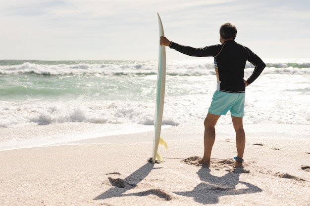 Вид сзади в полный рост на двухрасового пожилого мужчину, держащего доску для серфинга и смотрящего на волны в море в солнечный день