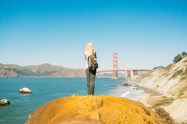 写真 明るい青い空に照らされたゴールデン・ゲート・ブリッジの海岸の岩の上に立っている女性の全長の後ろの景色