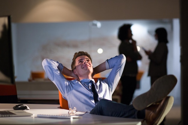 Full length of a relaxed casual young businessman sitting with\
legs on desk at night office