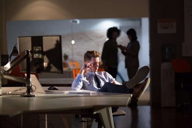 Full length of a relaxed casual young businessman sitting with\
legs on desk at night office