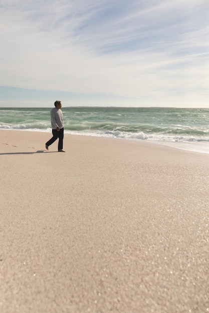 Full length of senior biracial man walking on shore at beach against sky during sunny day