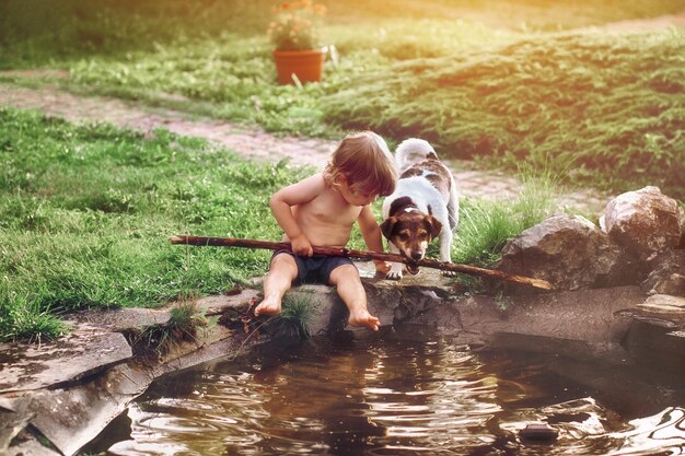 Photo full length of shirtless boy holding stick sitting by pond with dog