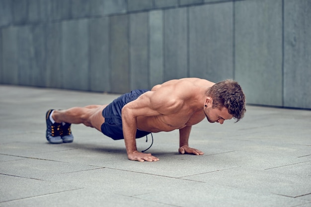 Full length of shirtless man doing push-ups on footpath by wall