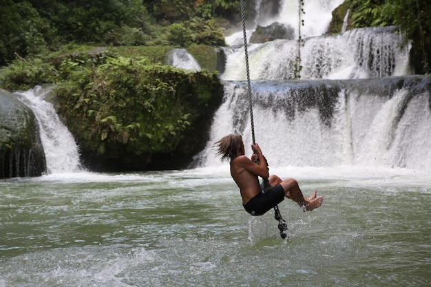 Foto lunghezza completa di uomo senza camicia appeso a una corda sul fiume contro la cascata