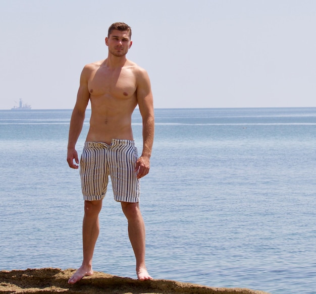 Full length of shirtless man standing on rock by sea against clear sky
