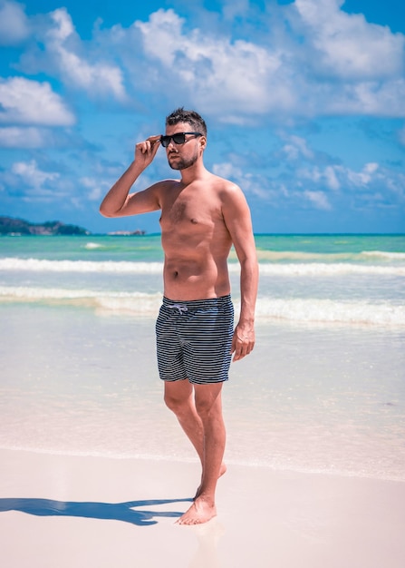 Full length of shirtless man in sunglasses walking at beach against sky