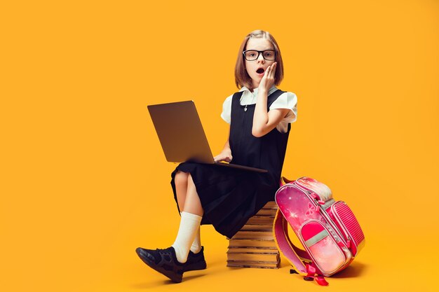 Шокированная школьница в полный рост сидит за стопкой книг с компьютером и смотрит в камеру образование детей