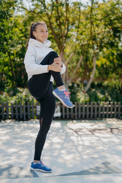 Full length shot of active healthy young woman warms up before jogging in park keeps knee raised wears sweatshirt leggings and sneakers listens music in earphones. Healthy lifestyle concept.