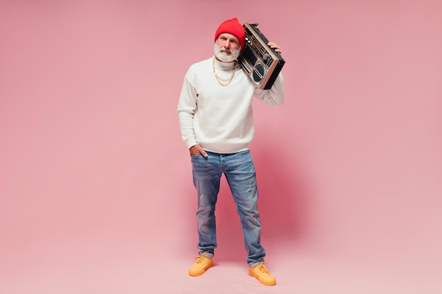 Full length shot of adult man with record player on pink background Fashionable guy with white beard in light sweater posing