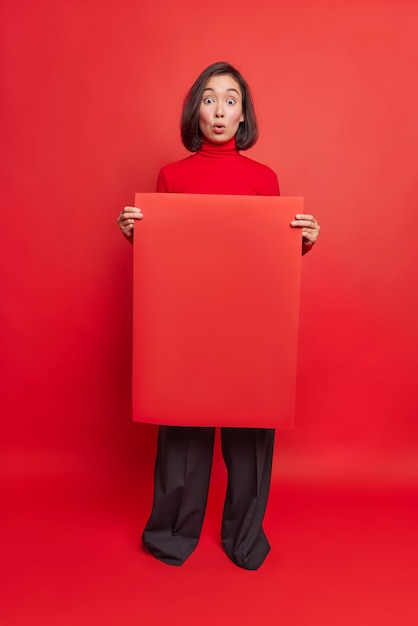 Full length shot of amazed Asian woman holds empty banner for your advertisement stunned by unexpected news wears turtleneck and black trousers isolated over red wall