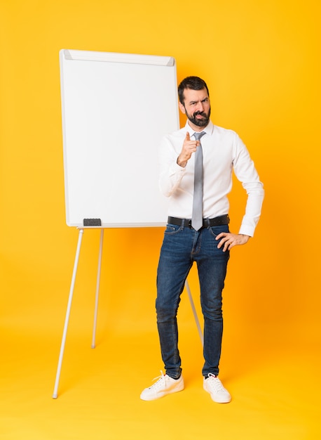 Full-length shot of businessman giving a presentation on white board over isolated yellow background frustrated and pointing to the front