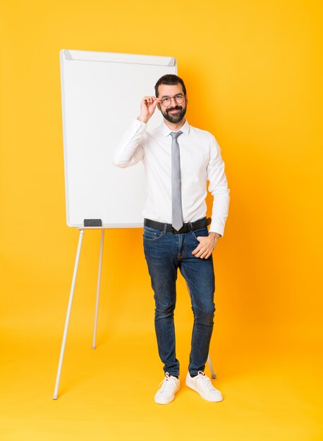Full-length shot of businessman giving a presentation on white board over isolated yellow with glasses and happy
