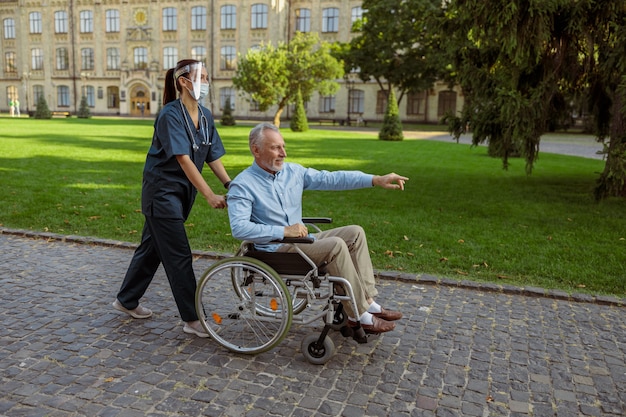 Full length shot of caring nurse wearing face shield and mask spending time with senior male