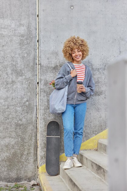 Full length shot of cheerful teenage girl drinks takeaway\
coffee stands on stairs against grey concrete wall rests after\
riding skateboard wears sweatshirt jeans and sportshoes has active\
lifestyle